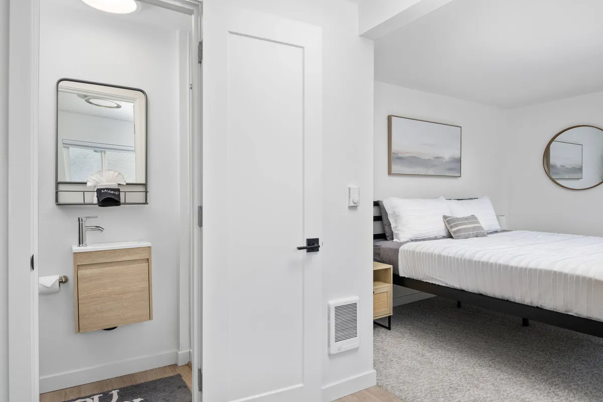 A serene bedroom interior featuring a king-size bed dressed in crisp white linens, a stylish vanity with a mirror, tasteful wall artwork, and a round mirror reflecting soft natural light. The scene exudes tranquility and comfort, with subtle ocean-inspired tones enhancing the restful mood.