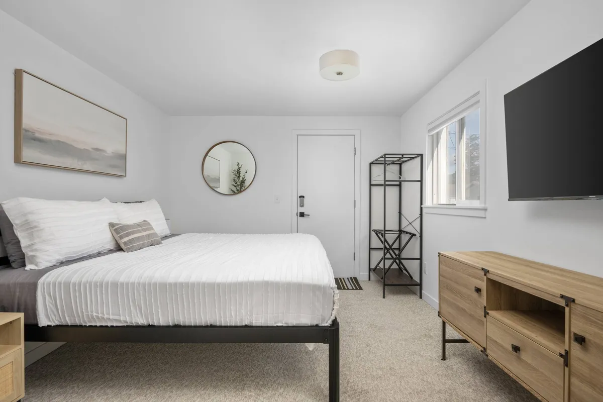 This inviting interior image centers on a plush bed flanked by a stylish mirror and wall-mounted TV, with coastal-inspired artwork adding a touch of local charm. The soft color palette and natural light create a soothing, peaceful ambiance perfect for unwinding after a day at the beach.