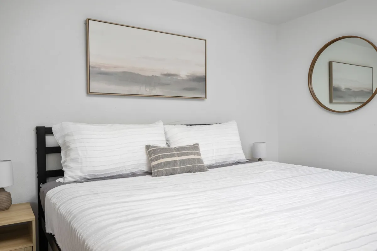 A cozy sleeping area with a comfortable bed, captivating artwork above the headboard, a nightstand with a reading lamp, and a mirror that reflects gentle, ambient light. The overall mood is quiet and intimate, ideal for unwinding or enjoying a quiet read.