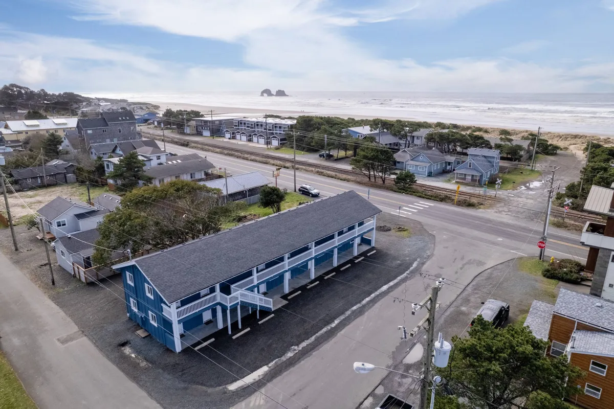 This exterior image features a blue beachfront inn with the ocean visible in the background and neighboring houses nearby. The setting exudes a relaxed beach town vibe, with people seen walking along the sandy shore and exploring local shops, suggesting easy access to both the beach and nearby amenities.