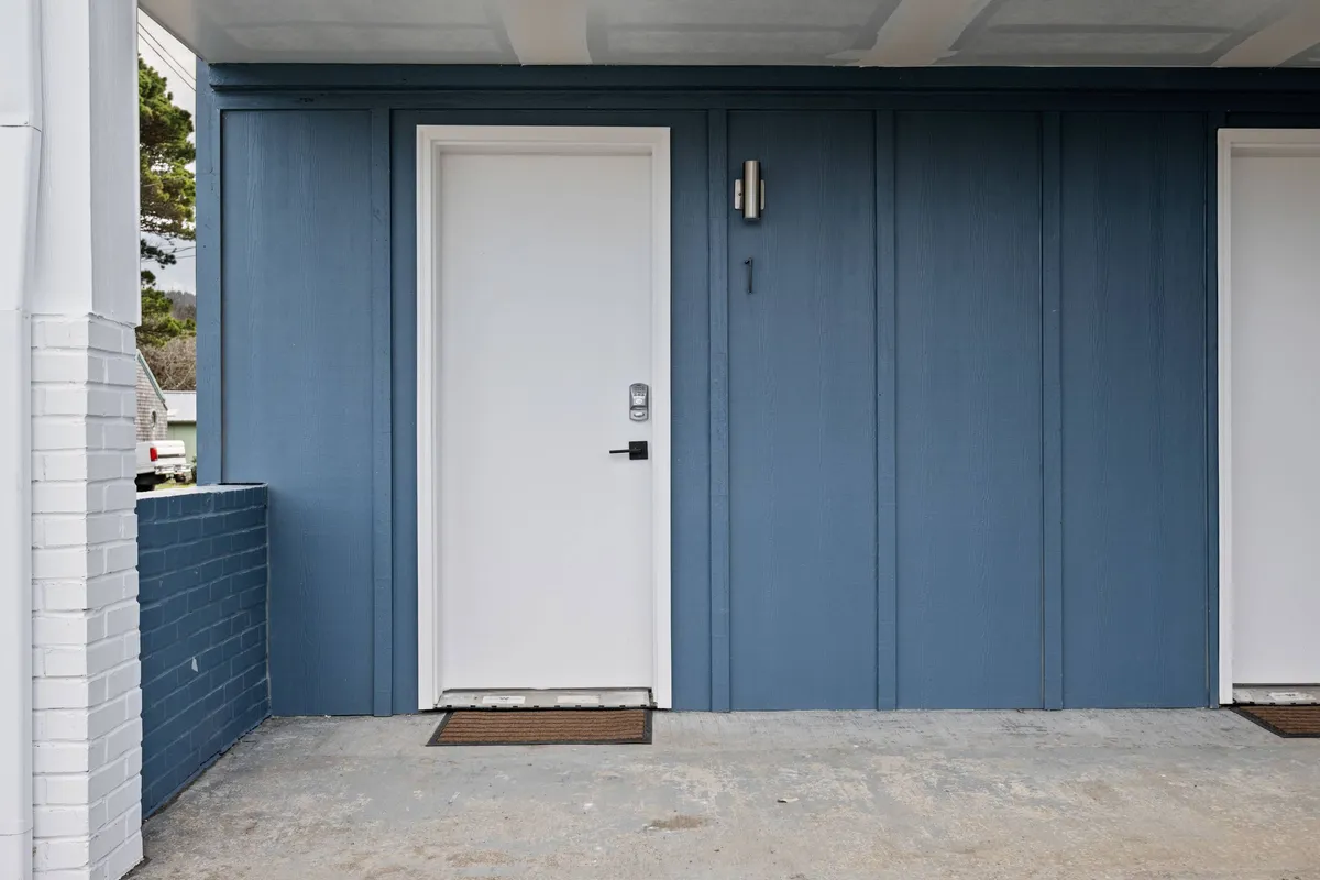 A bright, welcoming exterior view focusing on the blue wall, white door, and modern light fixture, with guests arriving or unloading luggage. The scene emphasizes a sense of arrival and comfort, suitable for both people and their pets.