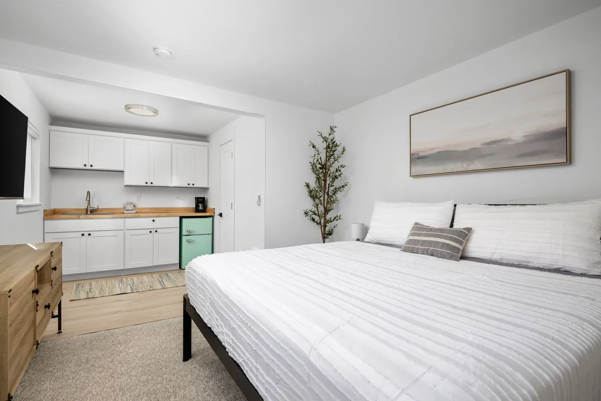 The image showcases a tranquil, stylishly remodeled condo interior featuring a plush bed with crisp white linens, a modern kitchenette highlighted by green accents, tasteful framed artwork, and a touch of greenery from an indoor plant. Soft natural light fills the space, enhancing the cozy and inviting atmosphere.