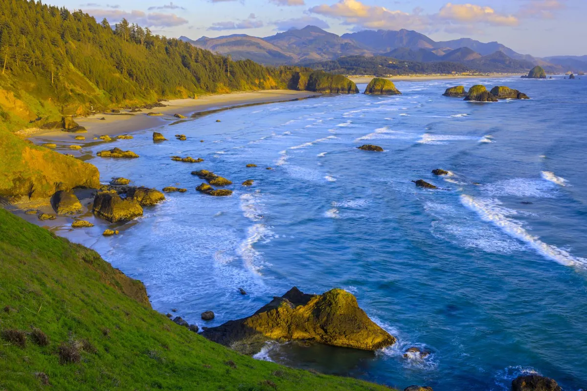 A wide, scenic view of the beach with ocean waves rolling in, dramatic rock formations, and distant mountains on the horizon. The shoreline invites leisurely walks, with opportunities for sunset watching, photography, and connecting with nature amid tranquil surroundings.